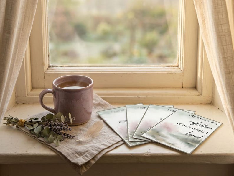 Lavender mug of tea on a linen napkin beside dried flowers, a feather and grief cards by a window